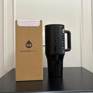 以下が含まれることがあります： ハンドルと黒い蓋が付いた黒いHydroJugが、段ボール箱の隣に置かれています。箱は茶色で、HydroJugのロゴとテキストが印刷されています。ジャグはテクスチャ加工された表面で、側面には「HYDROJUG」の文字が印刷されています。
