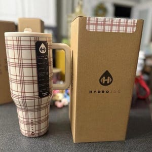 Może przedstawiać: Duży, izolowany kubek podr&oacute;żny z kremowo-brązowym wzorem w kratę. Kubek ma uchwyt i pokrywkę. Logo HydroJug jest widoczne na kubku i na dołączonym kartonowym pudełku.