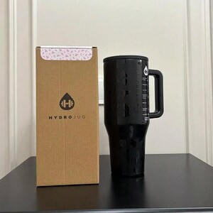 以下が含まれることがあります： ハンドルと測定マークが付いた黒いHydroJugウォーターボトル。HydroJugのロゴが入った茶色の段ボール箱の隣にあります。ボトルはテクスチャ加工された表面と黒い蓋が付いています。箱の上部には花柄があります。
