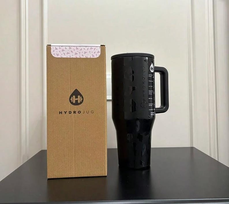 以下が含まれることがあります： ハンドルと測定マークが付いた黒いHydroJugウォーターボトル。HydroJugのロゴが入った茶色の段ボール箱の隣にあります。ボトルはテクスチャ加工された表面と黒い蓋が付いています。箱の上部には花柄があります。