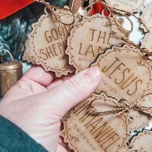 May include: Wooden Christmas ornaments with a rustic brown finish and gold string. Each ornament features a different Bible verse and phrase, including "Good Shepherd", "The Lamb", "Jesus", "Immanuel", and "Creator".