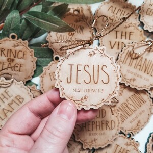 May include: A collection of wooden ornaments with religious text and scripture references. The ornaments are decorated with floral designs and have a rustic, natural look. The text on the ornaments includes "Jesus", "The Word", "Good Shepherd", "Cornerstone", "Prince of Peace", "Immanuel", "Alpha and Omega", "Author of Salvation", and "The Lamb".