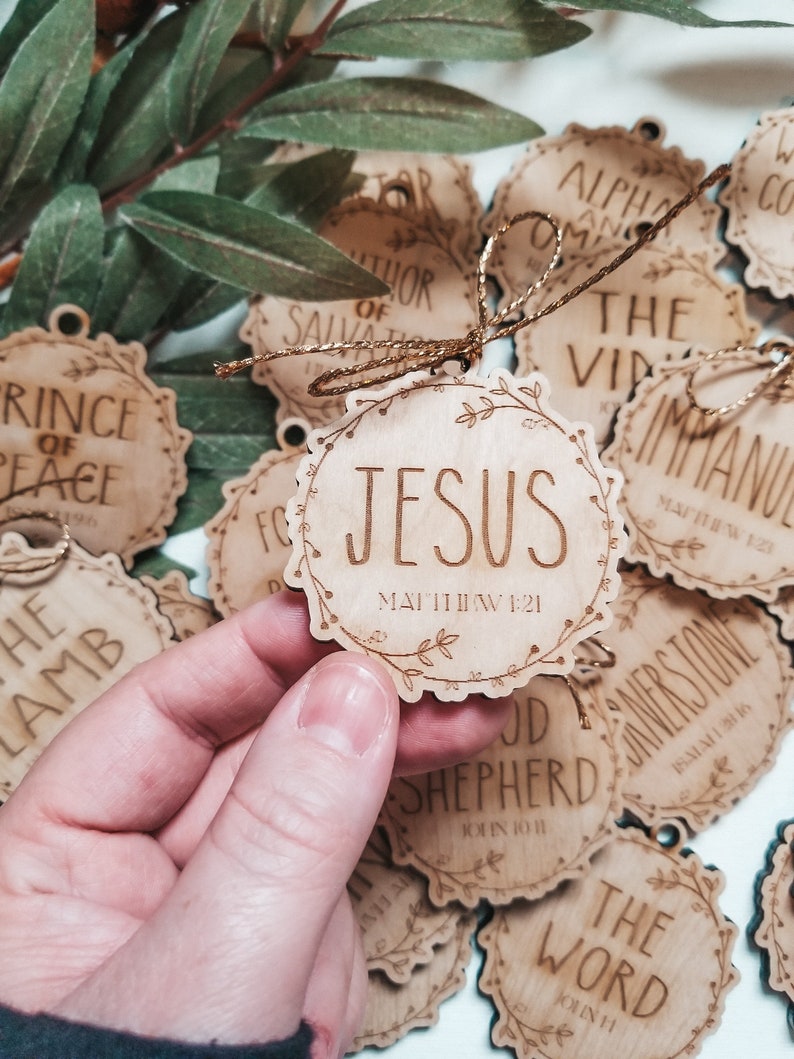 May include: A collection of wooden ornaments with religious text and scripture references. The ornaments are decorated with floral designs and have a rustic, natural look. The text on the ornaments includes "Jesus", "The Word", "Good Shepherd", "Cornerstone", "Prince of Peace", "Immanuel", "Alpha and Omega", "Author of Salvation", and "The Lamb".