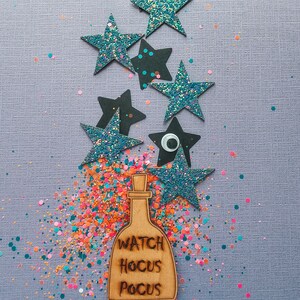 Peut inclure: Un charme de bouteille en bois avec le texte "Watch Hocus Pocus" entour&eacute; d'&eacute;toiles scintillantes bleues et de confettis color&eacute;s sur un fond violet.