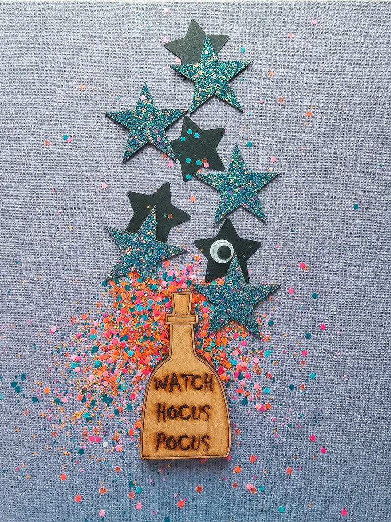 Peut inclure: Un charme de bouteille en bois avec le texte "Watch Hocus Pocus" entour&eacute; d'&eacute;toiles scintillantes bleues et de confettis color&eacute;s sur un fond violet.