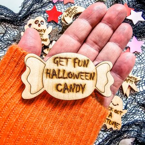Peut inclure: Une d&eacute;coration en bois en forme de bonbon avec le texte "Get Fun Halloween Candy" dessus. La d&eacute;coration est tenue dans une main portant un gant en tricot orange.