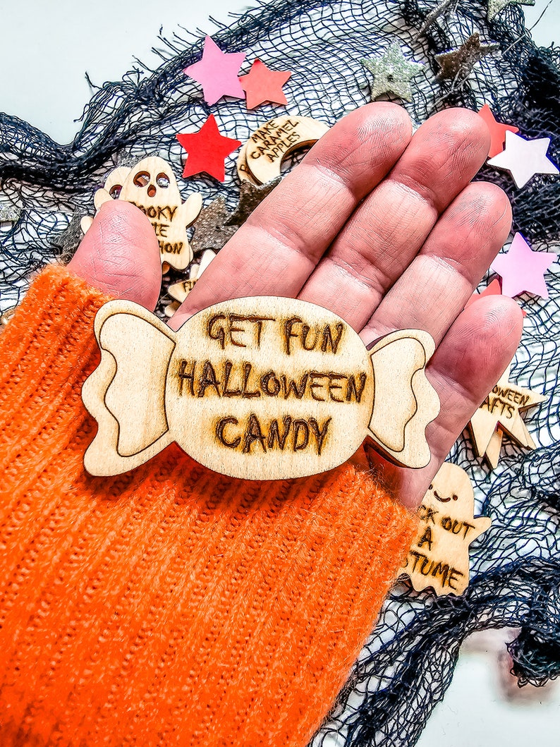 Peut inclure: Une d&eacute;coration en bois en forme de bonbon avec le texte "Get Fun Halloween Candy" dessus. La d&eacute;coration est tenue dans une main portant un gant en tricot orange.
