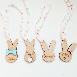 May include: Four wooden bunny-shaped ornaments with names "Luke", "Cooper", "Jameson", and "Mila" engraved on them. Each bunny has a different embellishment: a blue bow tie, a white pom-pom, a heart cutout, and a pink bow.