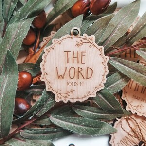May include: Wooden ornament with a floral wreath design and the words "The Word" and "John 1:1" engraved on it.