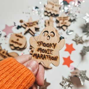 Peut inclure: Une d&eacute;coration en bois en forme de fant&ocirc;me avec le texte "Spooky Movie Marathon" &eacute;crit dessus. La d&eacute;coration est tenue dans une main portant un pull orange. L'arri&egrave;re-plan est une surface blanche avec des paillettes et d'autres d&eacute;corations en bois &eacute;parpill&eacute;es.