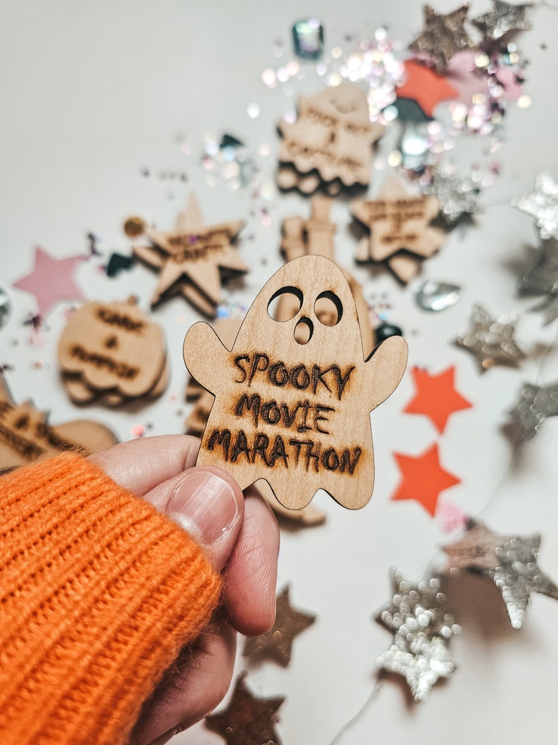 Peut inclure: Une d&eacute;coration en bois en forme de fant&ocirc;me avec le texte "Spooky Movie Marathon" &eacute;crit dessus. La d&eacute;coration est tenue dans une main portant un pull orange. L'arri&egrave;re-plan est une surface blanche avec des paillettes et d'autres d&eacute;corations en bois &eacute;parpill&eacute;es.