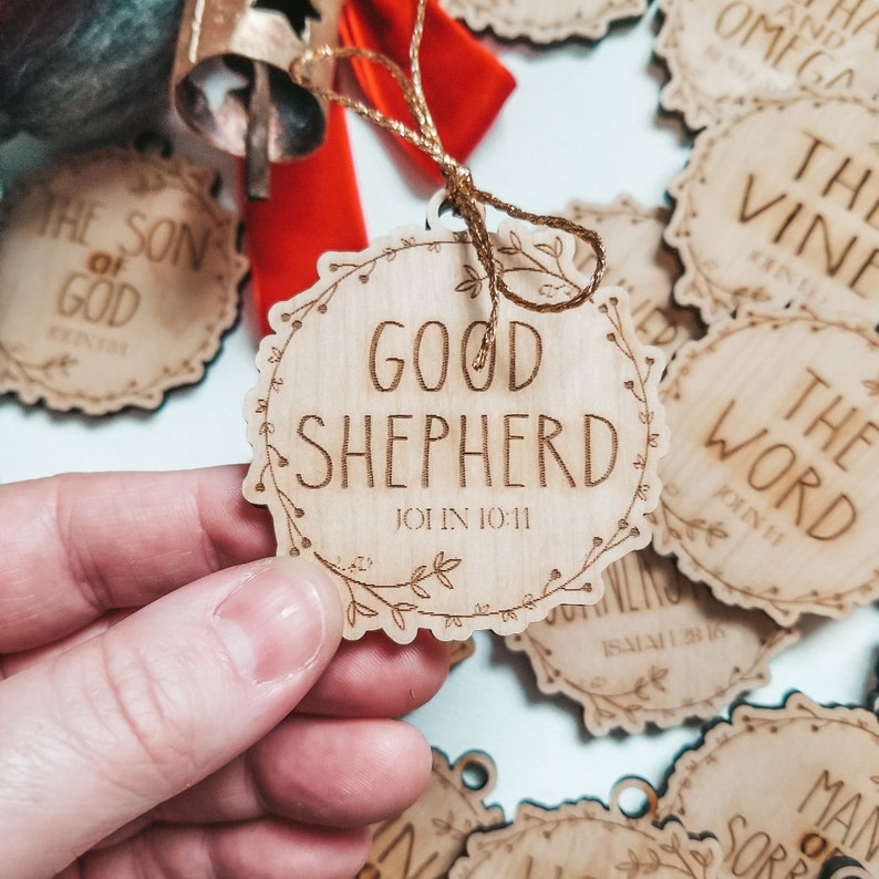 May include: A wooden ornament with the words "Good Shepherd" and the scripture reference "John 10:11" engraved on it. The ornament is decorated with a floral design and has a string attached for hanging.