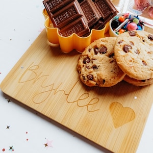 May include: A personalized bamboo serving board with the name "Brylee" engraved on it. The board has a heart shape engraved in the corner. There are chocolate bars, cookies, and sprinkles on the board.