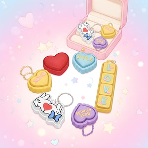 May include: A selection of heart-shaped and rectangular keyrings in various colours, including yellow, red, blue, and purple. Some keyrings feature text like "XOXO," "SMILE," and "Best Friends." A pink jewellery box holds a few keyrings. The background is a soft pink with heart and star shapes.