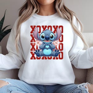 May include: White sweatshirt featuring a blue cartoon character with large ears, holding a heart shape with its hands. The design includes red "XOXO" text. The sweatshirt is a casual style.