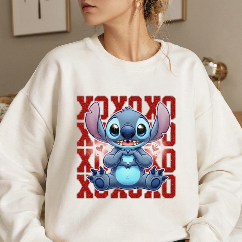 May include: White sweatshirt featuring a blue cartoon character making a heart shape with its hands. The design includes the text "XOXO" in red glitter letters. The character has large eyes and ears.