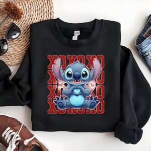 May include: Black sweatshirt featuring a blue cartoon character making a heart shape with its hands. The character is set against a red background with the text "XOXO" repeated vertically. The sweatshirt is a crew neck.