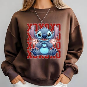 May include: A brown sweatshirt featuring a blue cartoon character making a heart shape with its hands. The character is set against a red glittery background with the text "XOXO" repeated. The sweatshirt has long sleeves and a crew neck.