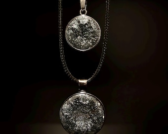 May include: Two silver-toned necklaces with round pendants. Each pendant features a textured, dark gray design encased in a silver frame. The necklaces hang against a black background, showcasing the jewelry's details.