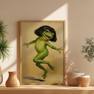 Puede incluir: Impresión artística enmarcada que presenta una rana verde con un corte de pelo bob negro, con los brazos extendidos y una expresión alegre. La rana está representada en una pose dinámica sobre un fondo cálido y neutro.