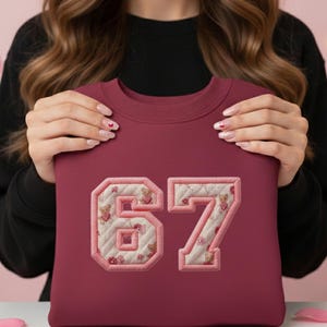 May include: Burgundy sweatshirt with the number "67" appliquéd on the front. The numbers are outlined in pink and filled with a quilted pattern of teddy bears and flowers. The sweatshirt is held up by a person.