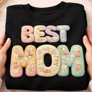 May include: Black sweatshirt with the words "BEST MOM" in pastel-colored, floral-decorated letters. The letters are in a 3D style, with pink, yellow, and blue hues. The sweatshirt is held by hands.