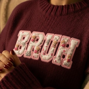 Pode incluir: Camisola de malha bordô com a palavra "BRUH" aplicada no peito. As letras são brancas com um padrão floral e acabamento rosa, com pequenos detalhes de ursinhos e corações. A camisola tem gola e punhos canelados.