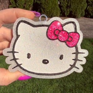 Puede incluir: Llavero brillante con la cara de un gato de dibujos animados con un lazo rosa brillante. La cara del gato es blanca con contornos y detalles negros. Un anillo plateado está unido en la parte superior.