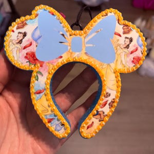 May include: A mouse-ear shaped ornament featuring a light blue bow at the top center. The surface is decorated with a repeating pattern of a female character in a yellow dress, red roses, and books. The edges are lined with small yellow beads.