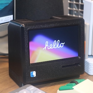 Puede incluir: Un monitor de computadora negro, compacto y de forma cuadrada con un acabado texturizado. La pantalla muestra la palabra hello en texto cursivo blanco sobre un fondo colorido y borroso. Un pequeño icono cuadrado azul y blanco está en la esquina inferior.