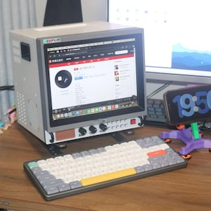 Könnte beinhalten: Ein Computer-Setup mit einem grauen Monitor, einer mechanischen Tastatur mit verschiedenfarbigen Tasten und einem Smartphone, das die Uhrzeit anzeigt. Das Setup befindet sich auf einem Holztisch.