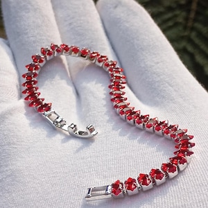 Può includere: Un braccialetto d'argento con una serie di vivaci pietre preziose rosse. Il braccialetto è appoggiato su una superficie bianca, e la chiusura è aperta. Le pietre preziose sono rettangolari e ravvicinate, creando una linea continua di colore. Il braccialetto è un gioiello.