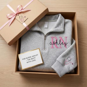 May include: A light gray quarter-zip sweatshirt with pink embroidered text reading RN and ashley, plus a pink stethoscope graphic on the sleeve. It is inside a cardboard gift box with a pink ribbon and a card that says FOR NURSE ASHLEY - THE BEST! WITH LOVE.