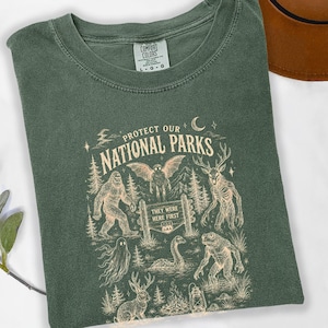 May include: A forest green t-shirt featuring a cream-coloured graphic with the text "PROTECT OUR NATIONAL PARKS" and "THEY WERE FIRST". The design includes illustrations of a sasquatch, a mothman, a deer-like creature, a ghost, and a lake monster.