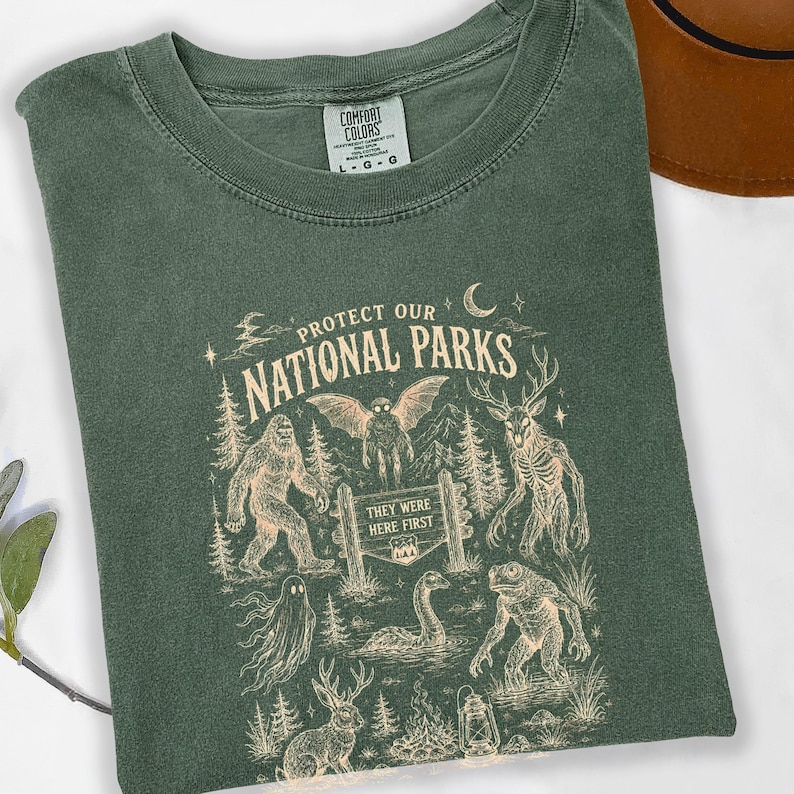 May include: A forest green t-shirt featuring a cream-coloured graphic with the text "PROTECT OUR NATIONAL PARKS" and "THEY WERE FIRST". The design includes illustrations of a sasquatch, a mothman, a deer-like creature, a ghost, and a lake monster.