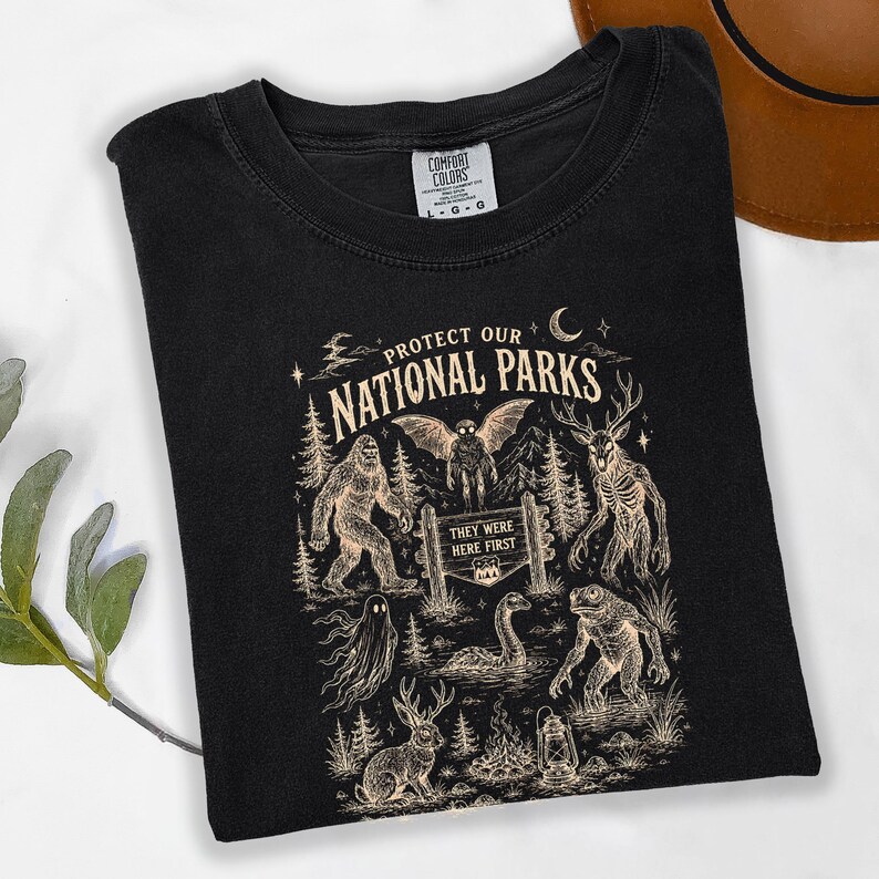 May include: A black crew neck t-shirt featuring a cream-coloured graphic print with the text "PROTECT OUR NATIONAL PARKS" and "THEY WERE HERE FIRST". The design includes illustrations of Bigfoot, a mothman, a deer-like creature, and a jackalope.