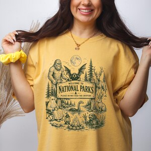 May include: A mustard yellow t-shirt featuring a dark green graphic print of a wooden sign that reads Welcome to National Parks Please Do Not Feed The Cryptids, surrounded by illustrations of Bigfoot, a mothman, a ghost, and other creatures.