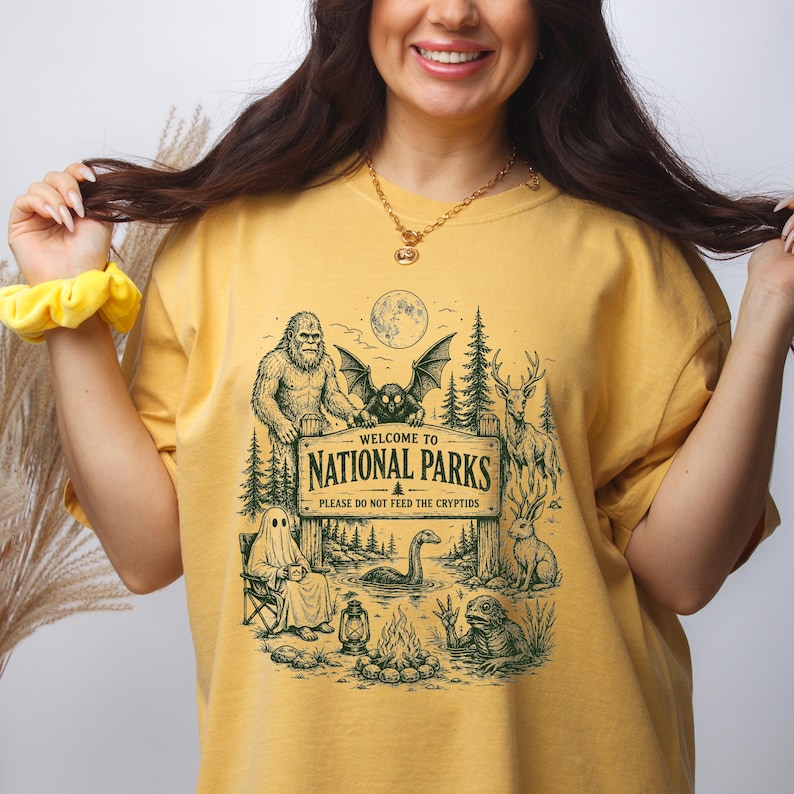 May include: A mustard yellow t-shirt featuring a dark green graphic print of a wooden sign that reads Welcome to National Parks Please Do Not Feed The Cryptids, surrounded by illustrations of Bigfoot, a mothman, a ghost, and other creatures.