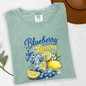 May include: A sage green t-shirt featuring a graphic print of a glass filled with yellow liquid, ice cubes, and blueberries. The text reads Blueberry Lemon. The design includes a lemon slice, a whole lemon, and a blue straw with a bow.