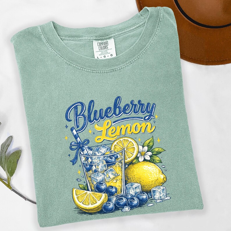 May include: A sage green t-shirt featuring a graphic print of a glass filled with yellow liquid, ice cubes, and blueberries. The text reads Blueberry Lemon. The design includes a lemon slice, a whole lemon, and a blue straw with a bow.