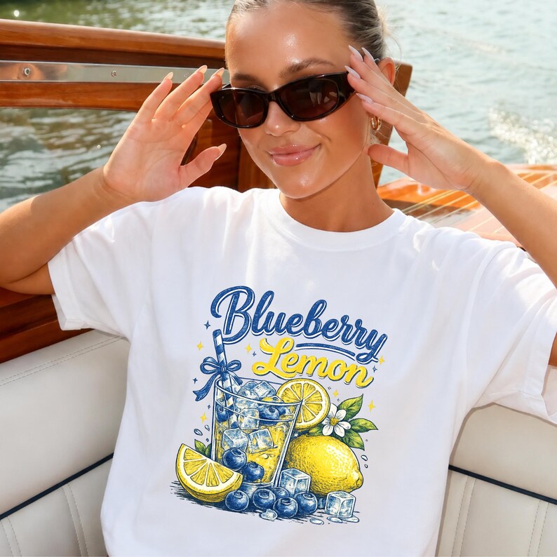 May include: A white t-shirt featuring a graphic print of a glass filled with a yellow lemon drink, ice cubes, and blueberries. The text "Blueberry Lemon" is printed in blue and yellow script above the glass, surrounded by lemon slices and leaves.
