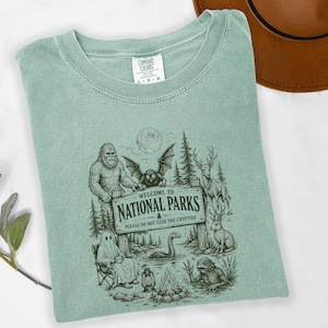 May include: A sage green t-shirt featuring a dark green graphic print of a sign that reads "WELCOME TO NATIONAL PARKS PLEASE DO NOT FEED THE CRYPTIDS" surrounded by illustrations of Bigfoot, a mothman, a ghost, a deer, a rabbit, and a lake monster.