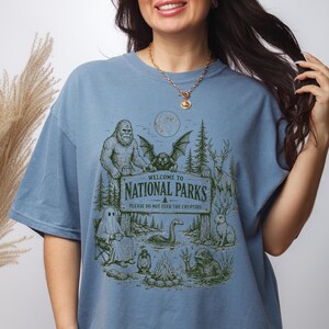 May include: A dusty blue t-shirt featuring a dark green graphic print of a National Parks sign that reads Welcome to National Parks, Please Do Not Feed the Cryptids. The design includes illustrations of Bigfoot, a ghost, a mothman, and a lake monster.