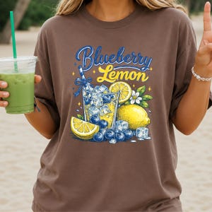 May include: A brown t-shirt featuring a graphic print of a glass filled with ice, blueberries, and lemon slices. The text "Blueberry Lemon" is printed in blue and yellow script above the illustration, which also includes a straw and small stars.
