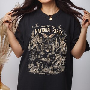 May include: A black t-shirt featuring a cream-coloured graphic print with the text "PROTECT OUR NATIONAL PARKS" and "THEY WERE HERE FIRST". The design includes illustrations of Bigfoot, a mothman, a deer-like creature, a ghost, a sea serpent, a frog-like creature, and a jackalope, along with trees, a moon, and a lantern.