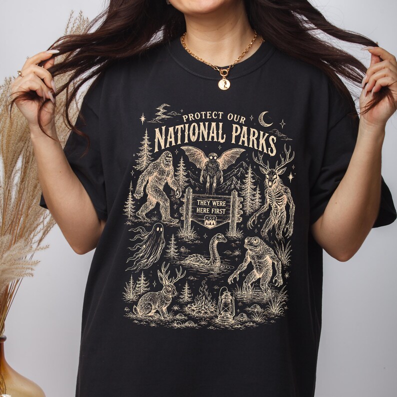 May include: A black t-shirt featuring a cream-coloured graphic print with the text "PROTECT OUR NATIONAL PARKS" and "THEY WERE HERE FIRST". The design includes illustrations of Bigfoot, a mothman, a deer-like creature, a ghost, a sea serpent, a frog-like creature, and a jackalope, along with trees, a moon, and a lantern.