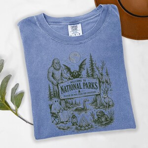 May include: A blue short-sleeve t-shirt featuring a graphic print of a sign that reads Welcome to National Parks Please Do Not Feed The Cryptids. The illustration includes Bigfoot, a mothman, a ghost, a deer, a jackalope, and a lake monster.