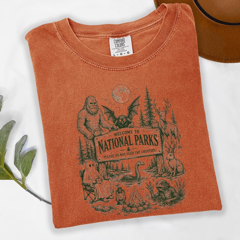 May include: A rust-coloured t-shirt featuring a dark green graphic of a wooden sign that reads "WELCOME TO NATIONAL PARKS PLEASE DO NOT FEED THE CRYPTIDS" surrounded by illustrations of Bigfoot, a mothman, a ghost, a deer, a jackalope, and a lake monster.