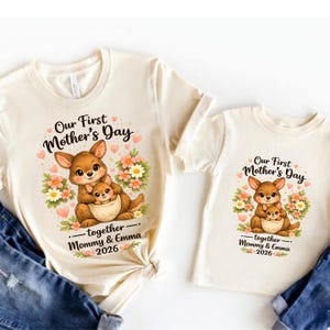 Może przedstawiać: Dwie kremowe koszulki z grafiką kangura. Grafika zawiera tekst "Our First Mother's Day" i "together - Mommy & Emma 2026". Kangur jest brązowy z małym kangurem w torbie, otoczony kwiatami.