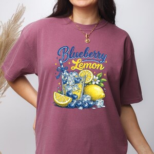 May include: A maroon short-sleeve t-shirt featuring a graphic print of a glass filled with ice, blueberries, and lemon slices. The text "Blueberry Lemon" is printed above the glass in blue and yellow script. The design includes small star accents.